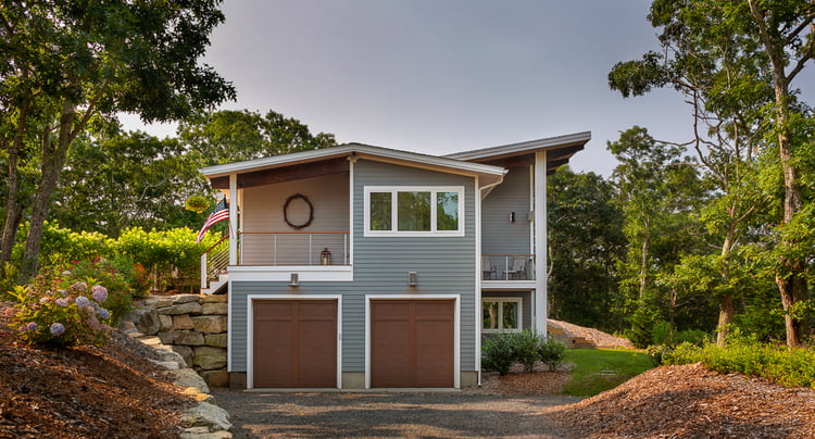 Truro Treehouse by Acorn Deck House Company is a modern farmhouse on Cape Cod. This photo shows the side exterior of the home, featuring gray siding, white windows, porch with cable railing, and two-car garage with brown doors.